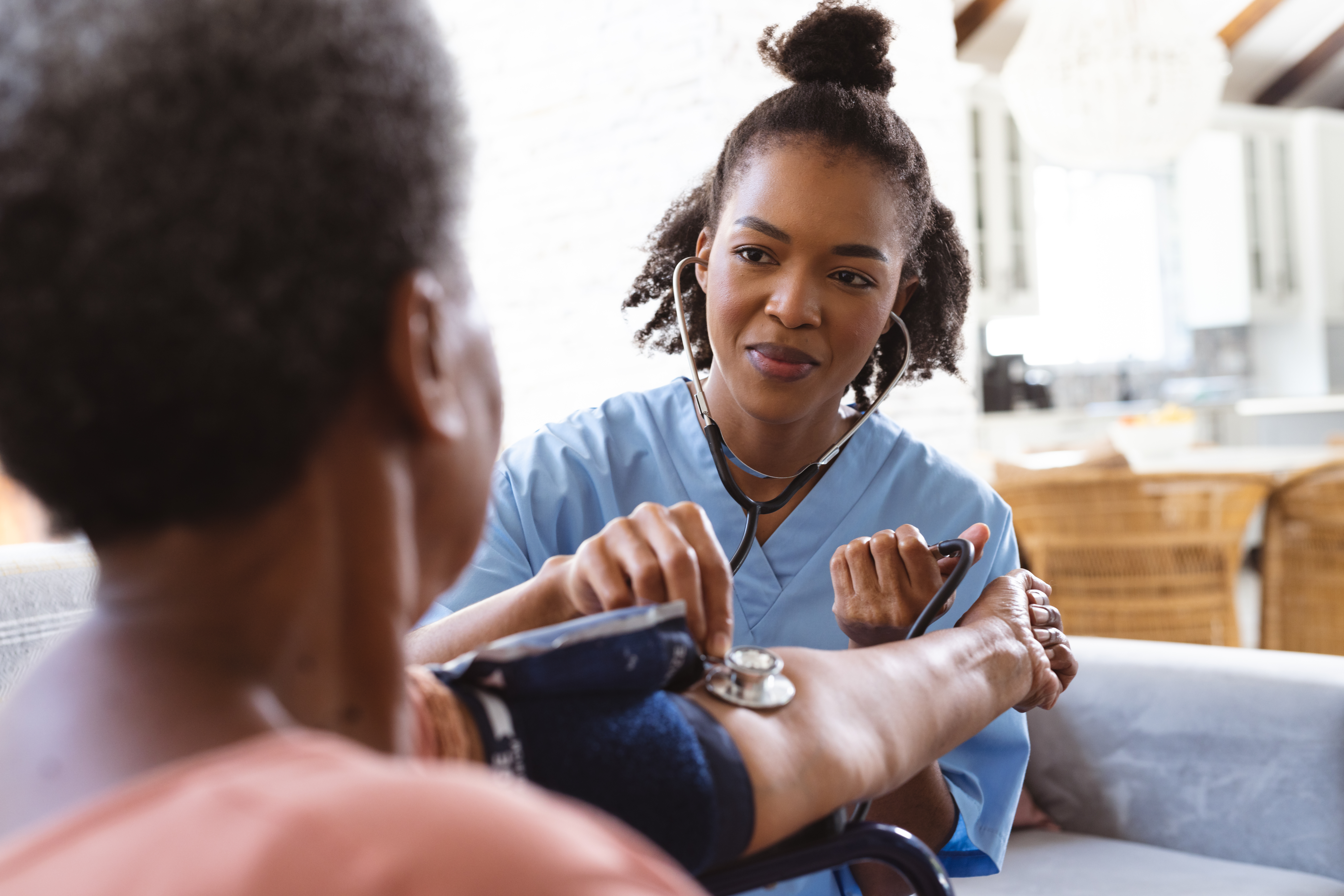 A nurse in blue scrubs checks a patient's blood pressure at home, conveying a caring and attentive atmosphere. The scene feels calm and professional.