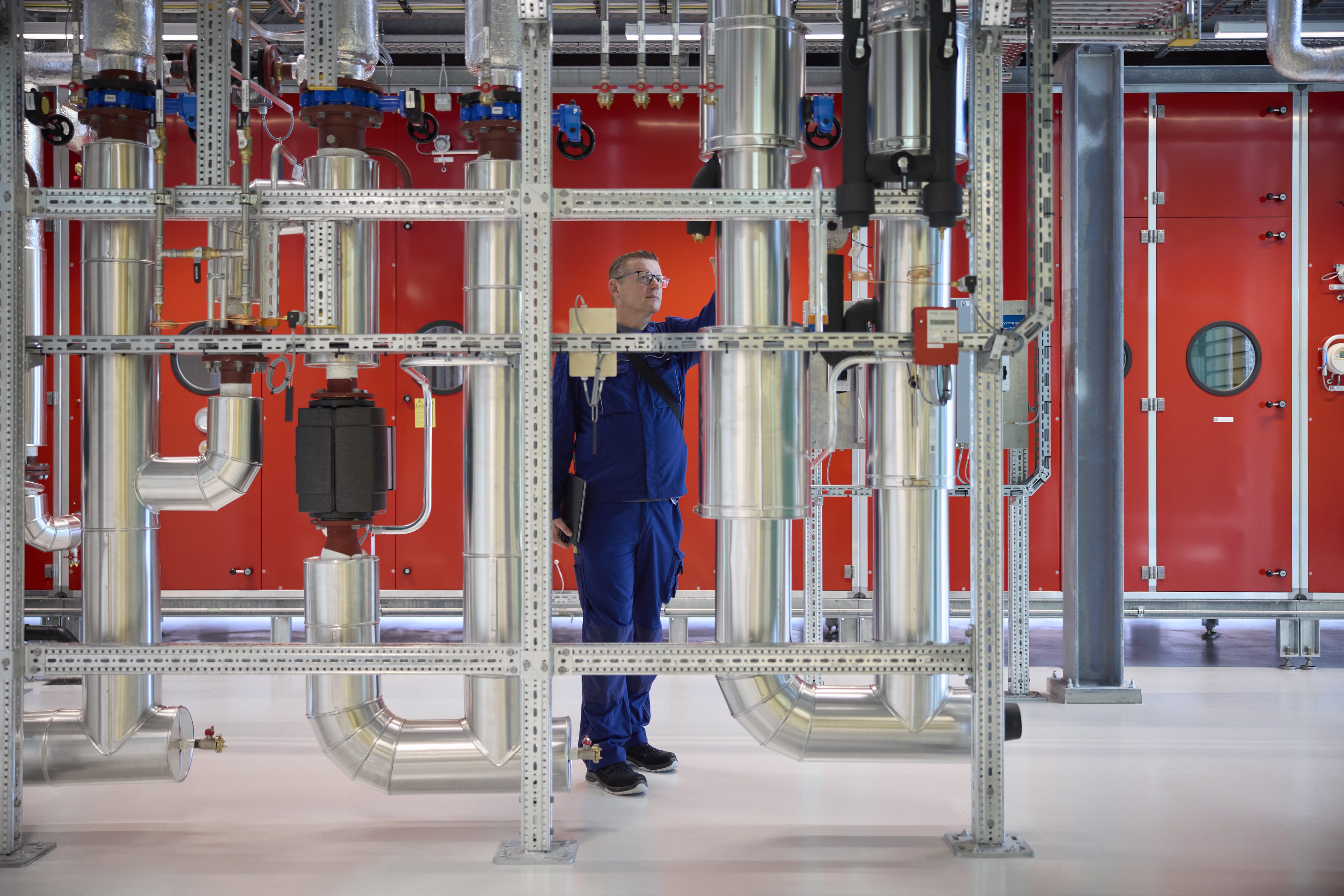 A technician in blue coveralls inspects industrial silver pipes and valves against a bright red wall. The setting is clean and organized.